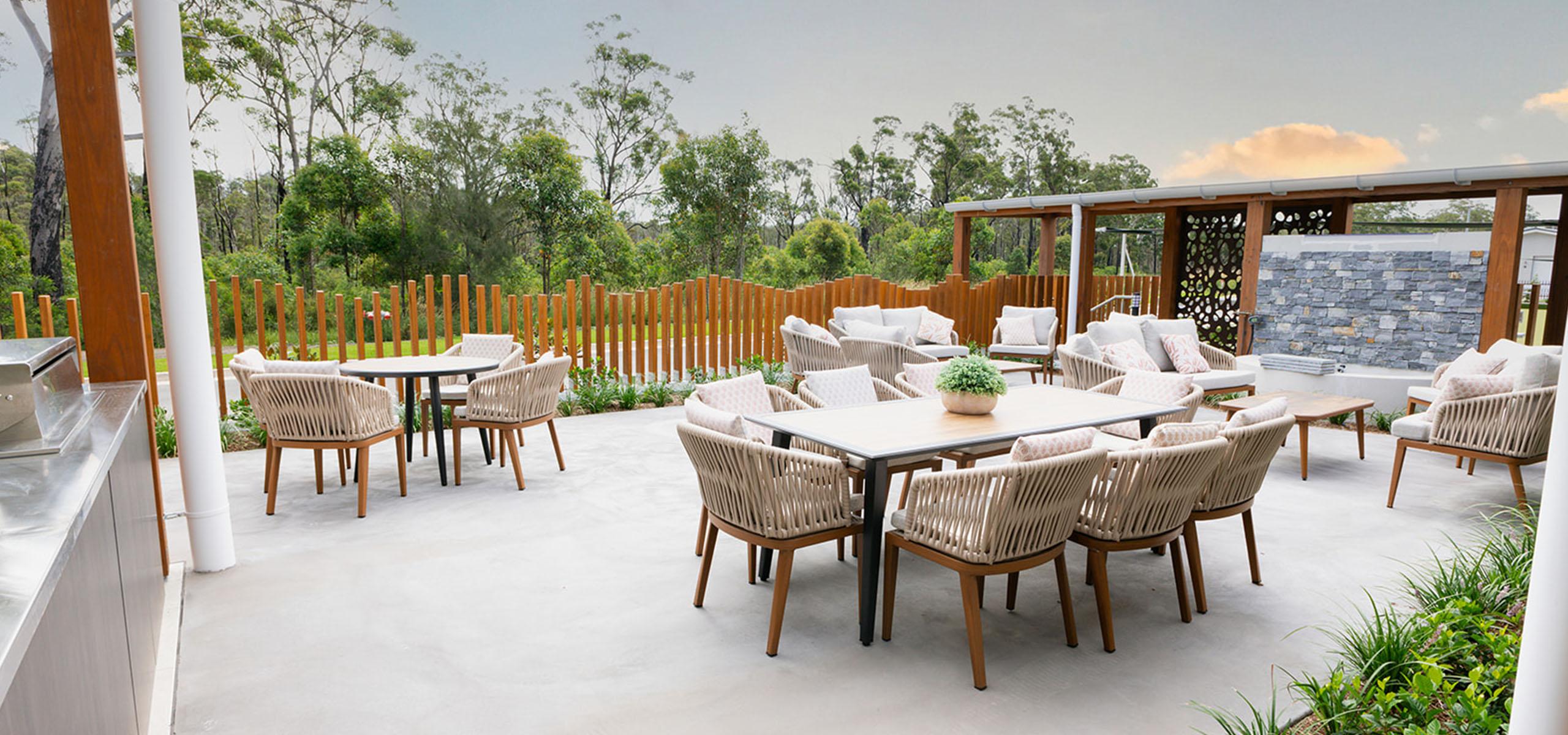 Stylish outdoor seating area in a lifestyle village with beige wicker chairs and tables on concrete patio, surrounded by greenery and wooden fence.