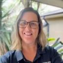 Person with glasses smiling in a black shirt, standing outdoors in a sunny, relaxed lifestyle village setting with green plants and a light-colored building.