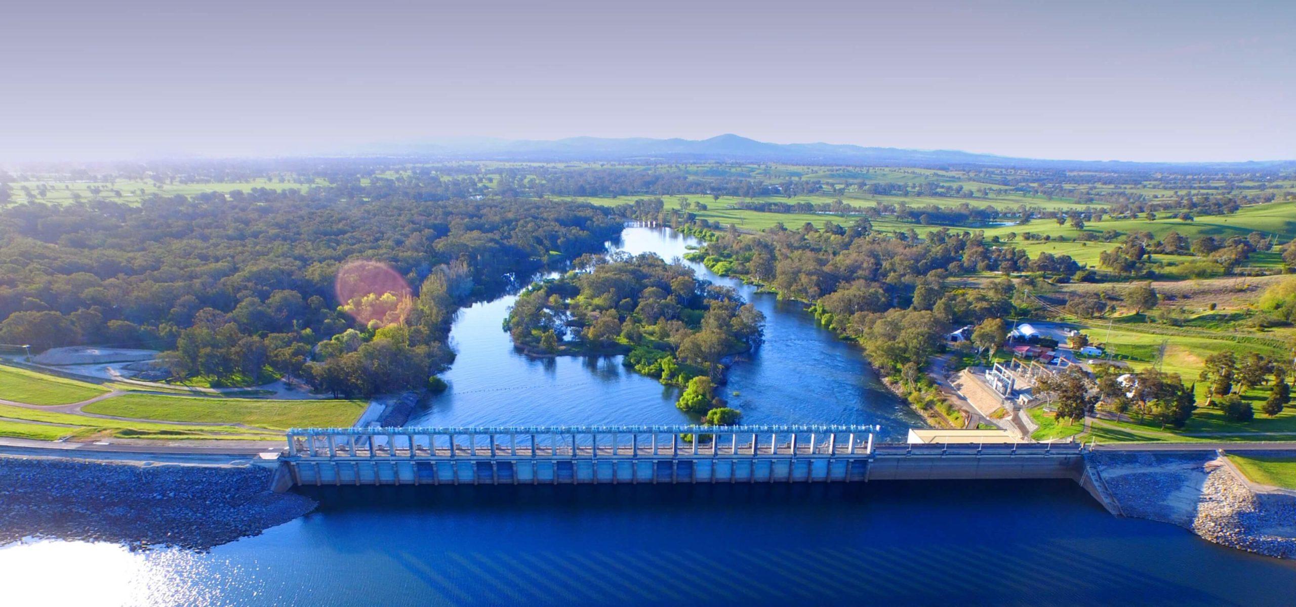 Dam spanning a river with water flowing underneath, surrounded by lush green fields, trees, and distant hills under bright, clear skies.