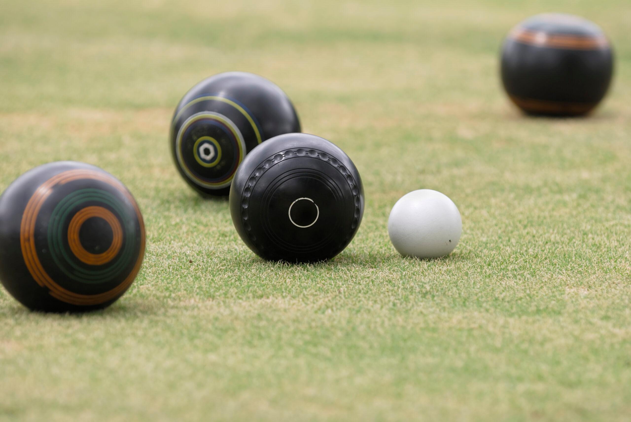 Four black lawn bowls and single white ball positioned on green grass surface, indicating game of lawn bowling in progress.
