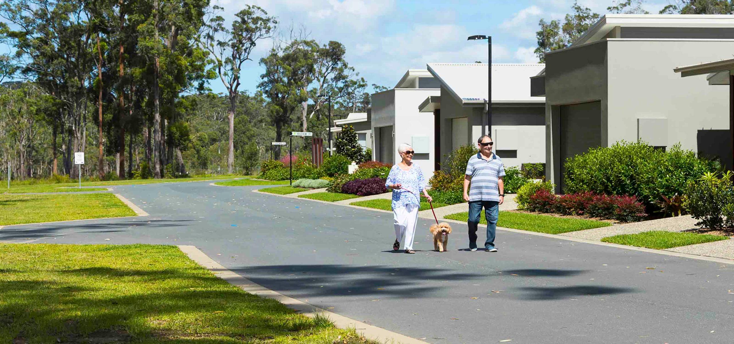 Elderly couple walking a small dog on a leash along a quiet suburban street in a lifestyle village, lined with modern homes and greenery under a blue sky.