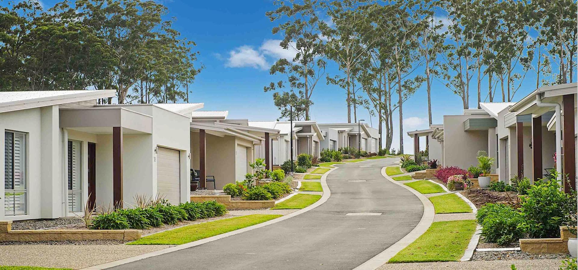 Row of modern lifestyle village houses with sloping roofs lining curved paved street, surrounded by landscaped gardens/tall eucalyptus trees under clear blue sky.