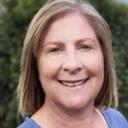 Woman (Lisa Fyle) with shoulder-length brown hair smiles warmly outdoors, green foliage background, suggesting friendly approachable lifestyle village atmosphere.