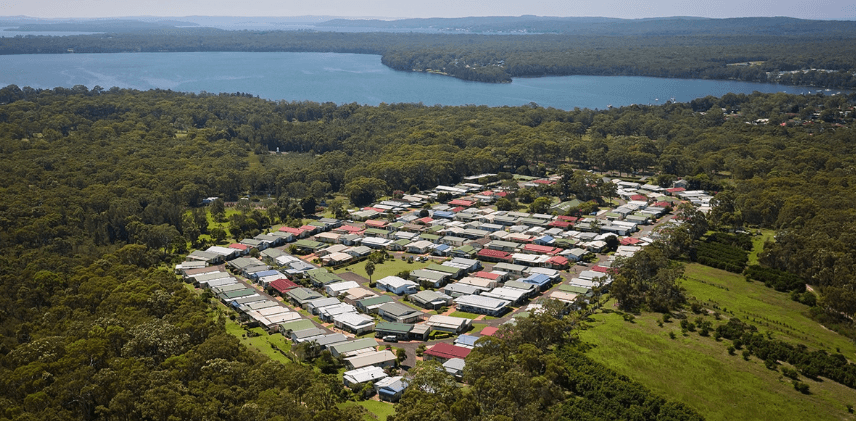 Aerial view of a lifestyle village residential area with houses on winding streets, surrounded by dense trees and a large body of water.