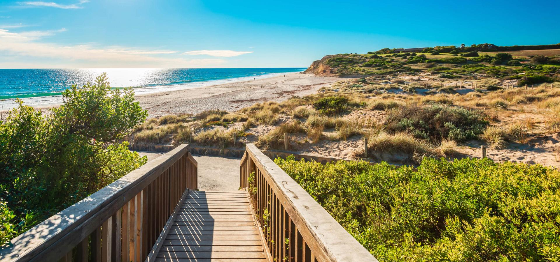 Wooden staircase leads down to sandy beach, calm ocean glistening under bright blue sky, lush greenery bordering path enhancing tranquil seaside atmosphere.