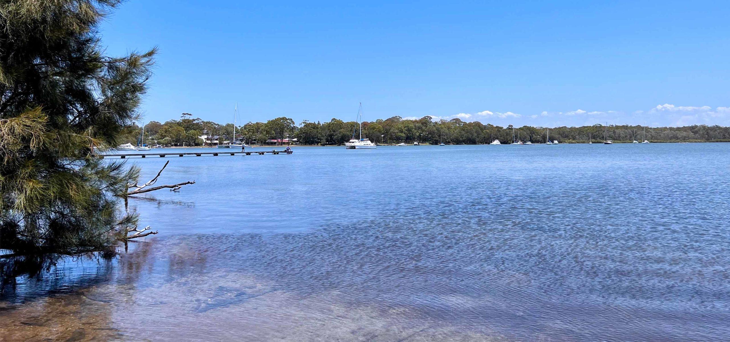 Calm lake reflecting a clear blue sky, with a wooden pier, anchored sailboats, and surrounded by the lush greenery of a lifestyle village environment.