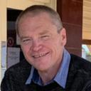 Smiling individual with short hair wearing dark sweater and light blue shirt in a casual setting, possibly outdoors in a lifestyle village.