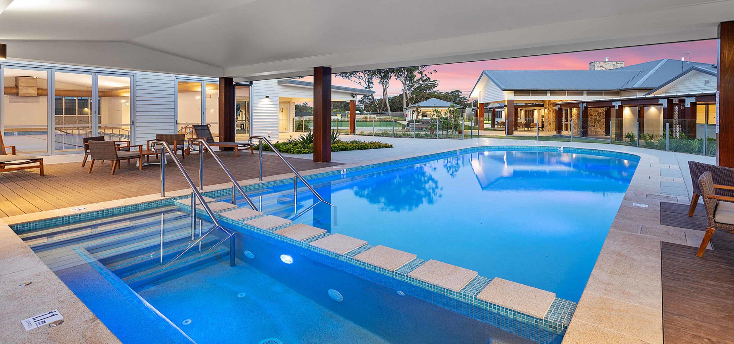 Lifestyle village swimming pool with steps/handrails reflecting sky, surrounded by wooden deck and cozy seating, nearby modern building with large windows.