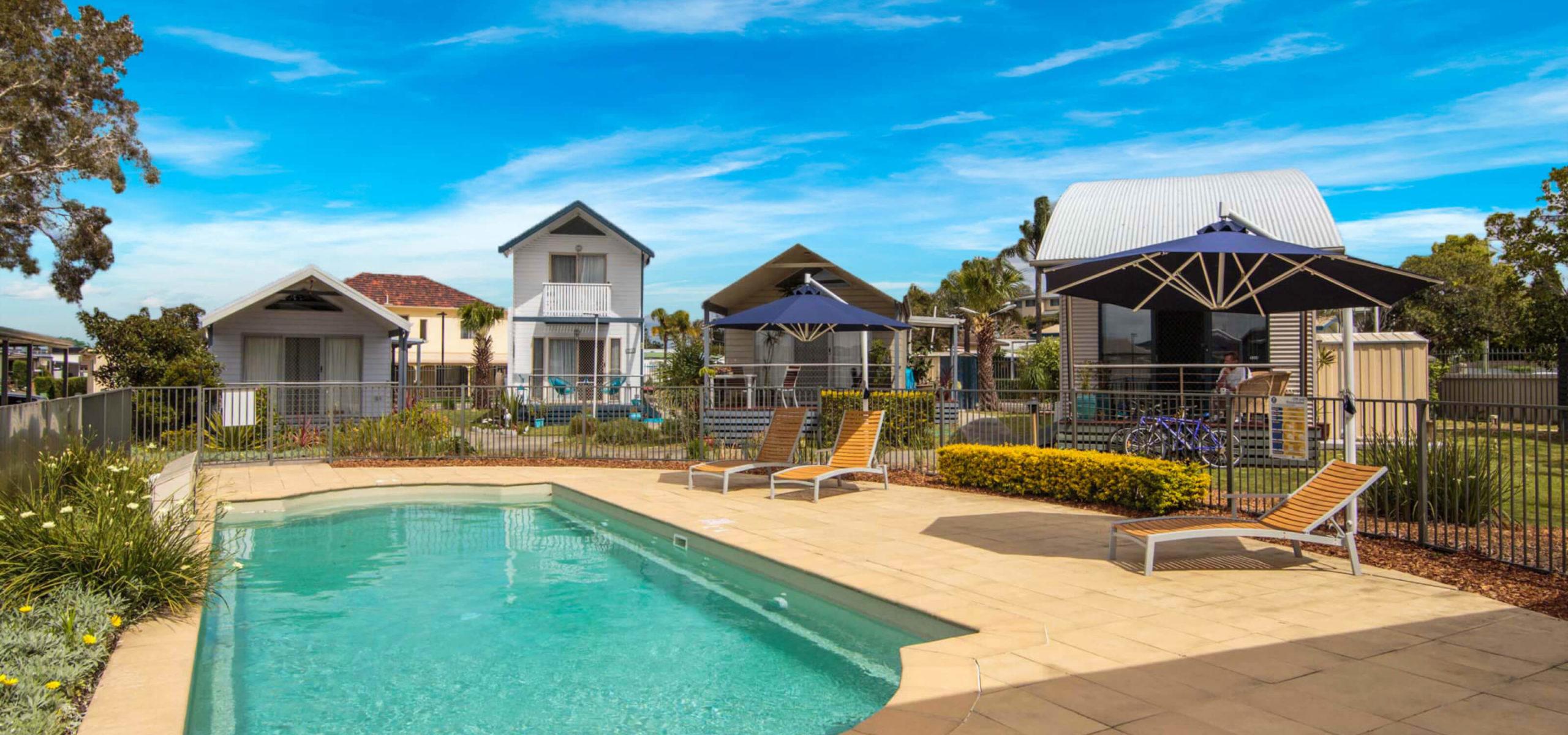 Lifestyle village swimming pool reflecting blue skies, surrounded by sun loungers, several beach-style houses flanked by greenery behind.